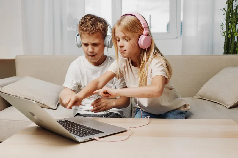 Two children with headphones interact with a laptop on a wooden table in a bright living room, focused on digital activity and learning.