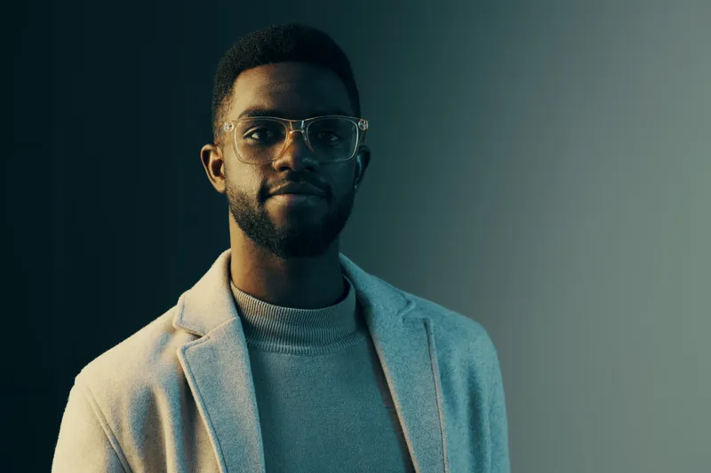 Stylish African man in glasses wearing a light gray coat, posing confidently against a modern gradient background, showcasing professionalism and sophistication