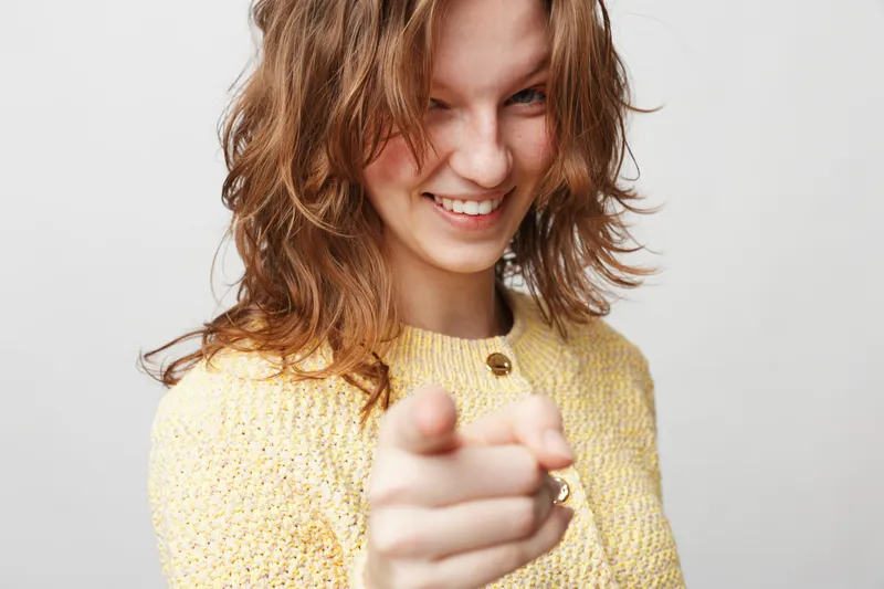 Smiling young woman with wavy brown hair wearing yellow sweater points finger at camera. Studio portrait on white background. Concept of confidence, friendliness, and positive attitude.