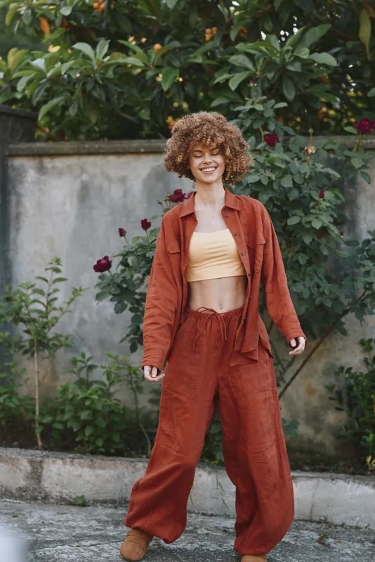 A cheerful woman outdoors wears a burnt orange suit with a pale yellow top, presenting casual fashion and a confident, sunny mood in a bright urban garden setting