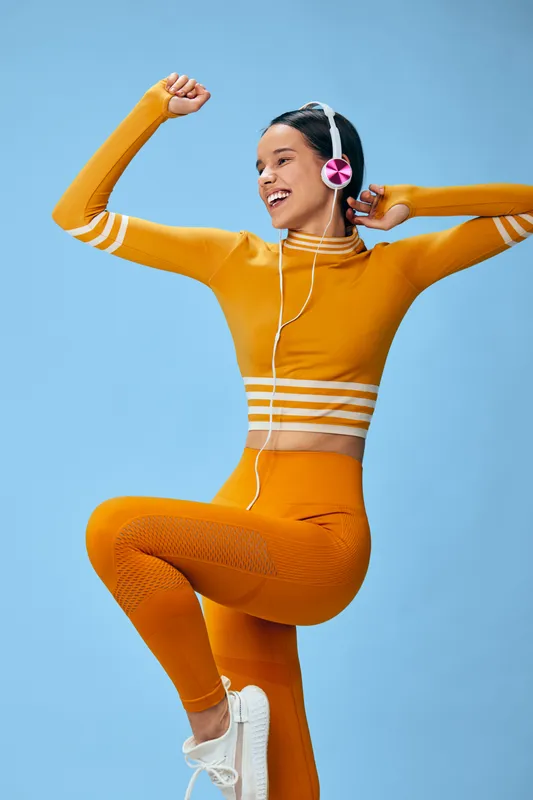 Woman fitness model in orange crop top and leggings performing energetic aerobic pose with raised knee and fists, wearing pink headphones against a clean studio blue background, joyful sport scene.
