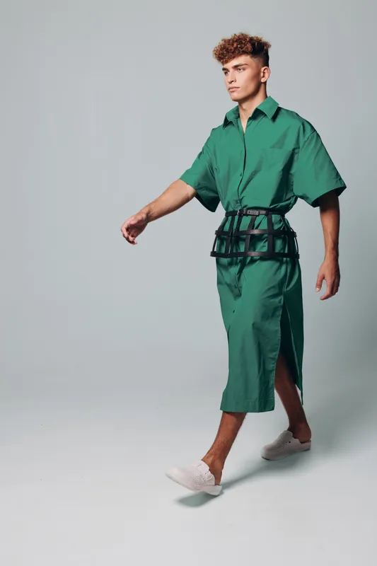 Young man walking in green oversized shirt and skirt with black belt accessory and white slip on shoes on gray background in studio.