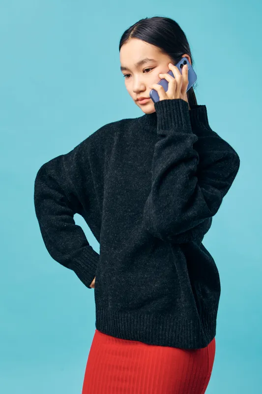 Young woman in black sweater and red skirt talking on smartphone against blue background with thoughtful expression and hand on hip for communication and technology.