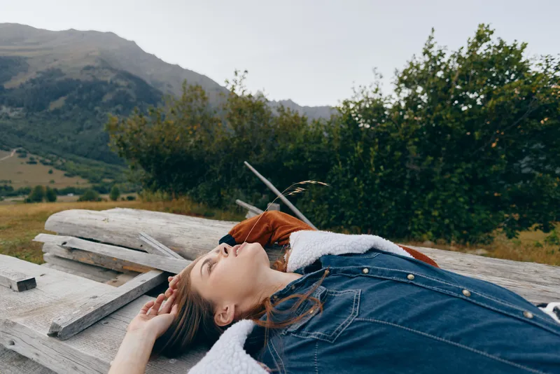 Woman lying on wooden bench in nature with mountains background, relaxed portrait in denim jacket outdoors, cozy layers and peaceful relaxation during countryside escape.