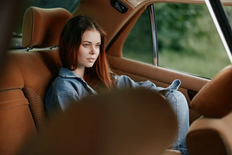 A thoughtful young woman with long brown hair sits in a cozy car interior, dressed in casual denim attire, reflecting on life amid a serene green backdrop.