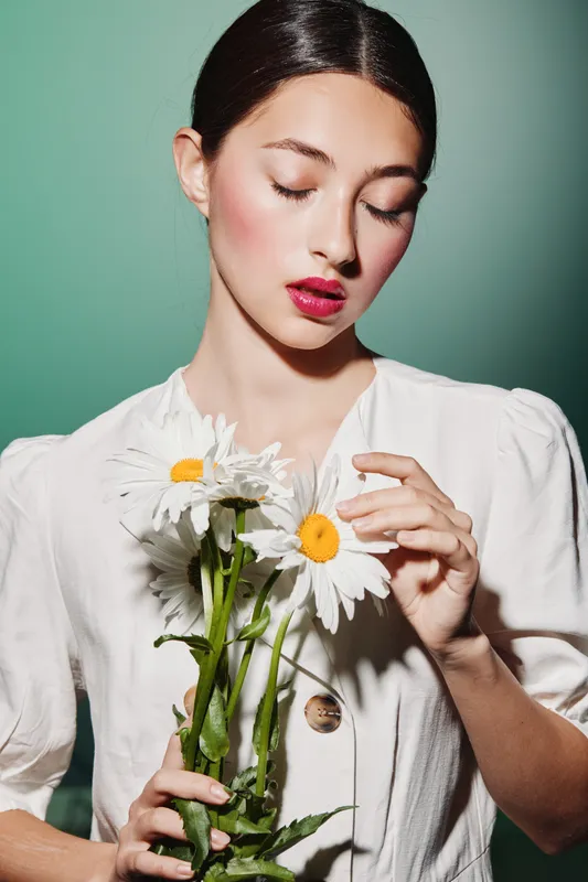 Young woman with bold red lipstick holding delicate daisies against a soft green background. She wears a white blouse with subtle texture. Concept for beauty, fashion, and creativity.