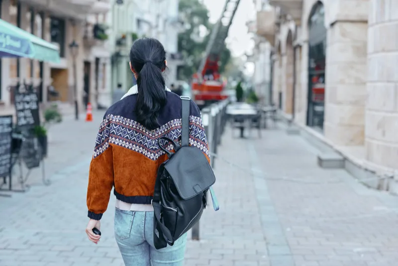 Backpack clad traveler walks away along a cobbled street in a casual urban setting, orange and navy patterned jacket, jeans, and calm mood of exploration and travel.