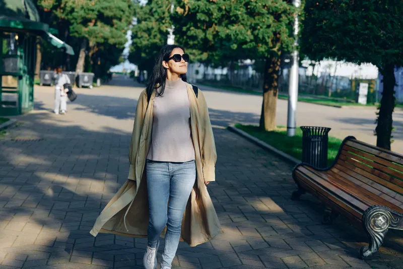 Woman wearing light trench coat and jeans walks along a sunny park street, sunglasses on, breeze flowing, urban nature mood, casual style, everyday street fashion moment