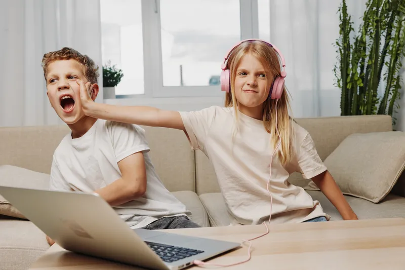 Two children at home on couch, girl wearing headphones playfully pushing boy's face, casual moment of sibling fun and interaction with laptop.