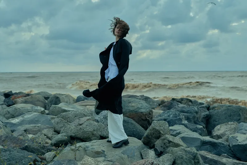 Sea, rocks, windy atmosphere with flowing coat and dress, figure standing on rugged shoreline, motion framing a dramatic outdoor moment, style and resilience amid ocean spray and gray skies
