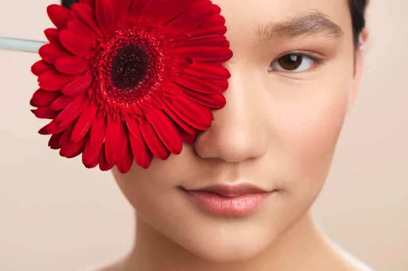 Asian woman with a vibrant red flower held beside her eye, presenting a soft, elegant portrait. The close frame emphasizes beauty, calm expression, and subtle artistry in a studio setting.