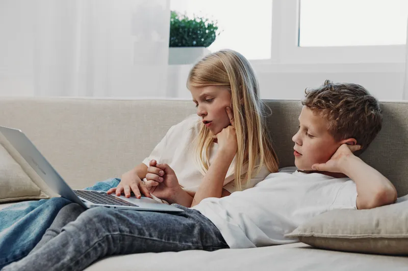Two children relaxing on a sofa using a laptop together, focused and engaged in a bright room with natural light. Concept of technology, learning, and childhood leisure time.