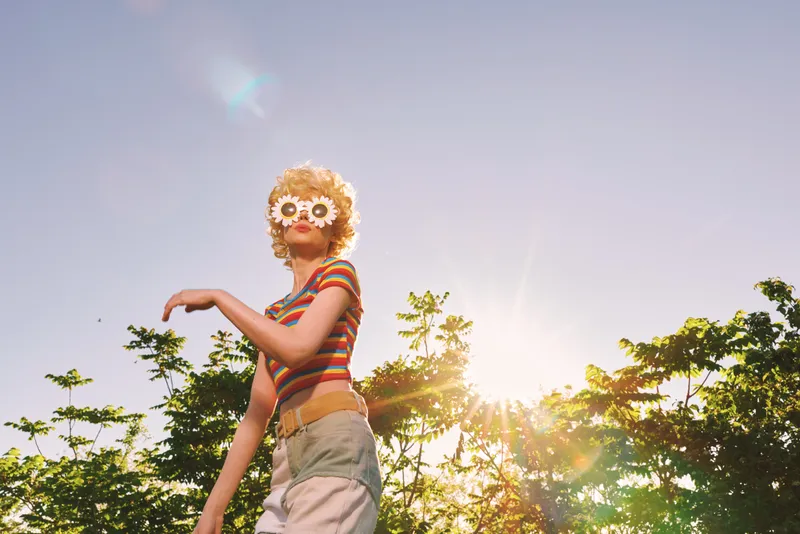 Young woman wearing striped top and floral glasses dances playfully outdoors as the sun flare and warm golden hour glow bathe her motion and smile.