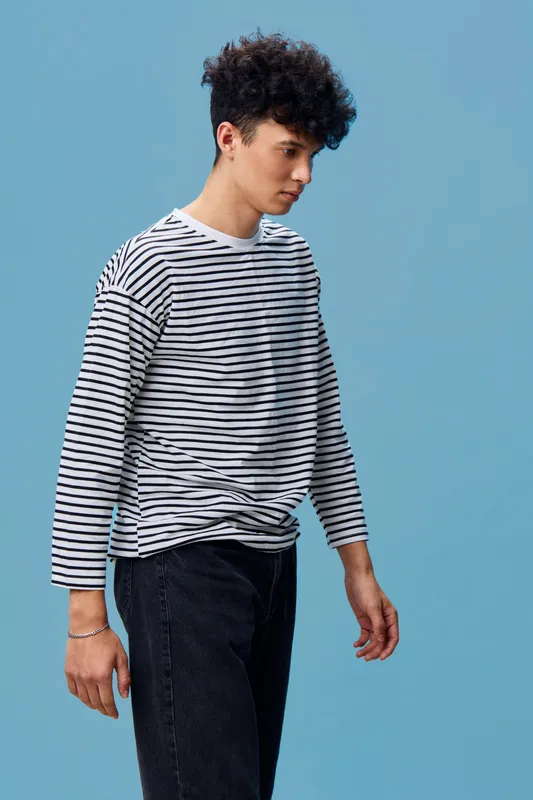 young man wearing striped long sleeve shirt and black jeans standing against blue background in casual style with serious expression and curly hair