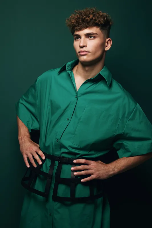 Young man with curly hair wearing oversized green shirt and black belt posing with hands on hips against dark green background in studio portrait.
