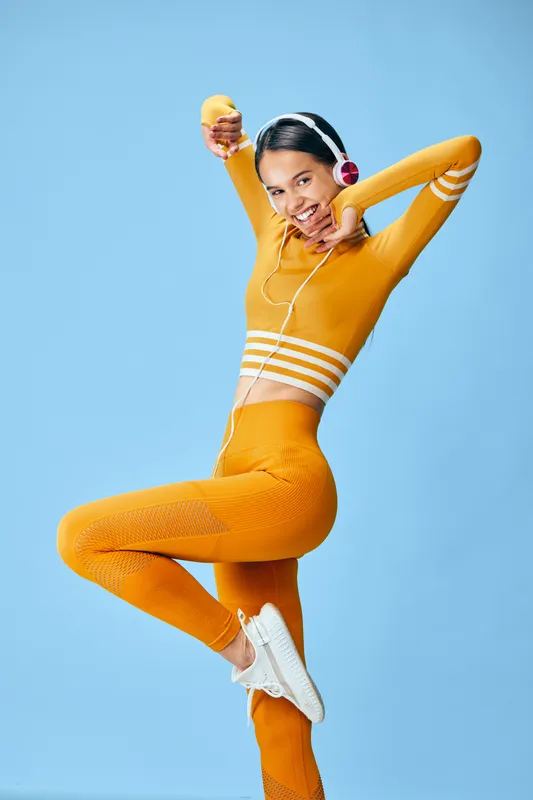 Energetic woman fitness model in bright orange workout outfit with crop top and textured leggings, wearing headphones and holding a water bottle in a dynamic studio pose on blue background.