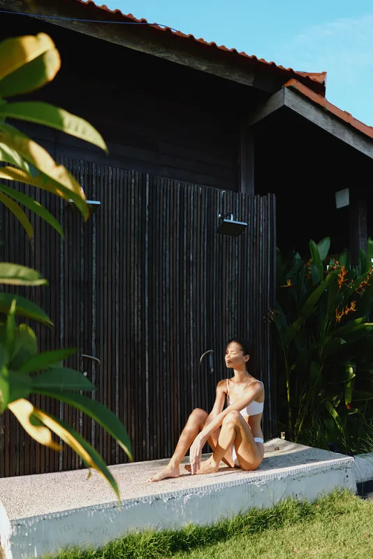 Woman outdoors, relaxed on a sunlit concrete ledge in a white swimsuit, warm breeze, modern dark wood backdrop, lush greenery and tropical plants, casual summer lifestyle moment, candid and peaceful