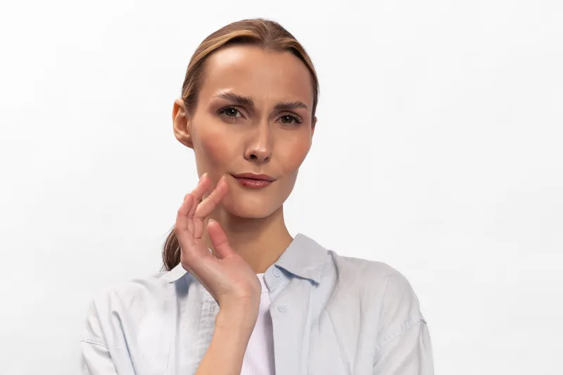 Thoughtful young woman with light brown hair in casual shirt looks at camera with questioning expression, isolated on white background. Concept for doubt, decision, and curiosity.