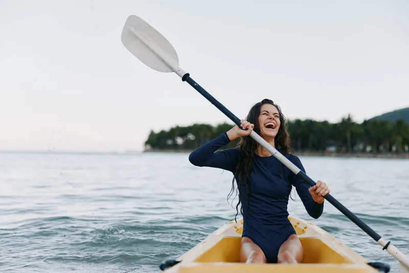 A joyful female paddles a yellow kayak across calm water, raising a paddle in a carefree pose as spray glints and distant shoreline frames a sunny scene