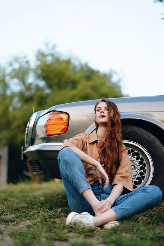 Young woman sitting beside a vintage car, wearing casual fashion and expressing contemplation in a natural outdoor setting.
