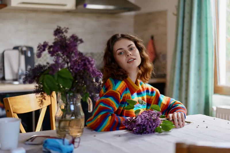 Cheerful woman indoors at a sunlit dining table, wearing a colorful striped sweater, with lilac bouquet, relaxed pose, cozy home moment, natural light, vibrant mood, casual elegance
