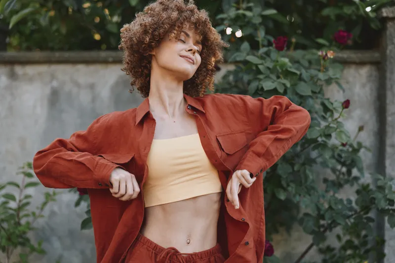 Young woman with curly hair wears an orange jacket over a pale yellow crop top in a garden setting, posing with hands on her hips and a confident, carefree vibe