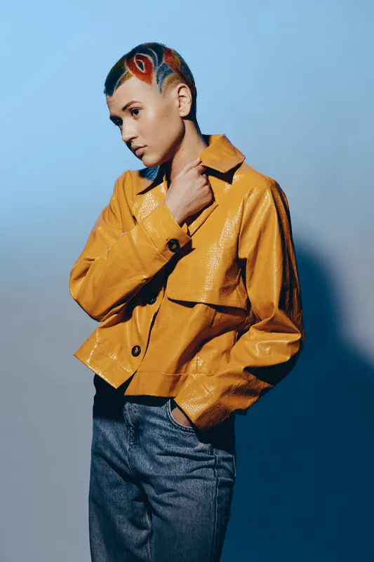 young woman with artistic shaved hairstyle wearing a glossy mustard jacket and denim pants, posing in dramatic studio lighting on blue-gray background