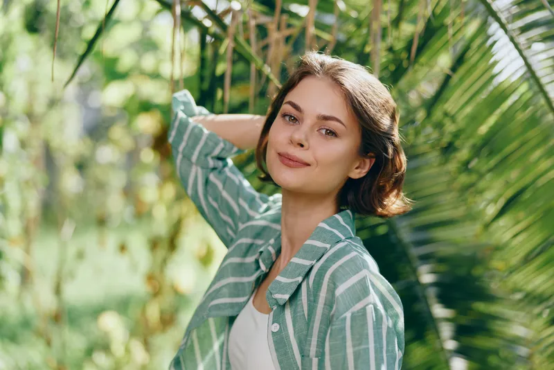 Portrait of a young woman outdoors, smiling softly, wearing a striped green shirt, surrounded by lush greenery.