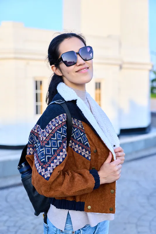Fashion forward outdoor scene with a casual stylish woman in a patterned jacket, scarf and backpack, wearing sunglasses, exuding urban vibe and confidence.