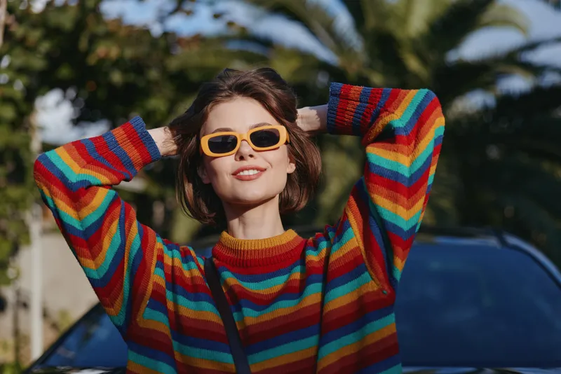 Woman in a colorful striped sweater wearing yellow sunglasses, smiling with hands behind head by palm trees and a car, casual outfit for travel lifestyle and sunny outdoor mood.
