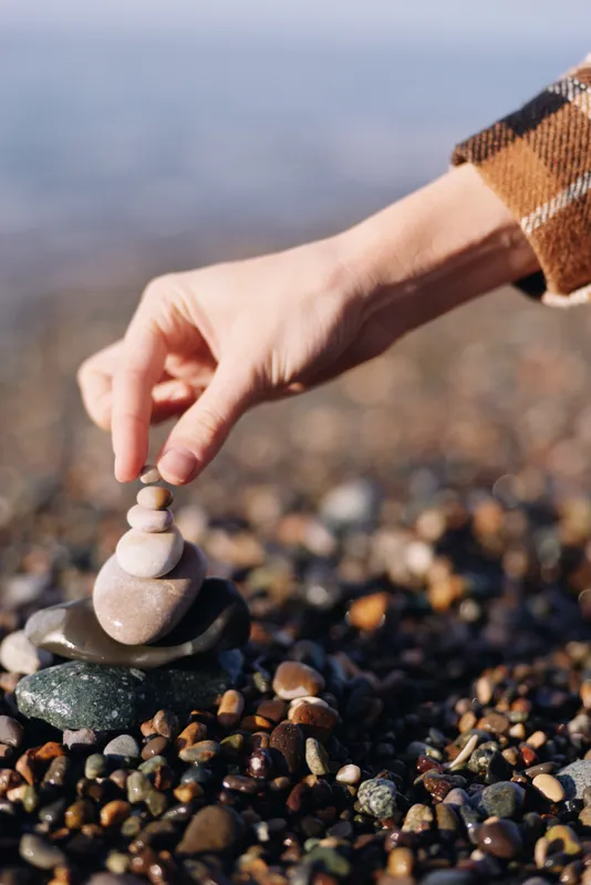 Hand places a small stone stack on a pebble beach, calm shoreline scene, mindfulness in action, simple balance and focus, natural texture, soft light and peaceful moment for relaxation