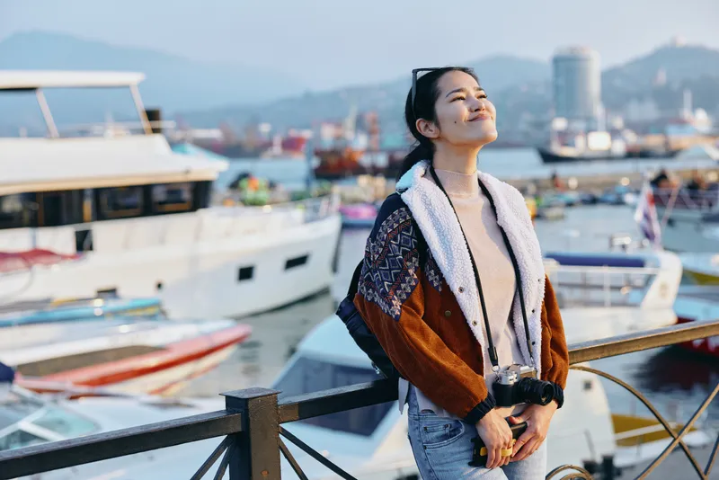 Smiling woman wearing a thick knit jacket on a harbor pier with boats and water in the background, showcasing fashion, travel, street style and a carefree outdoor moment