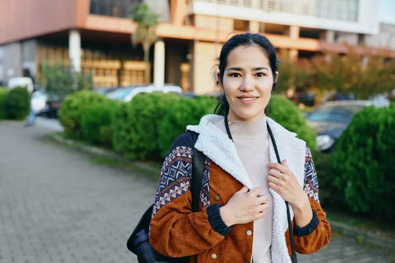 Woman wearing a brown patterned jacket and backpack on a city street, smiling and adjusting the zipper, urban casual style captured in natural daylight.