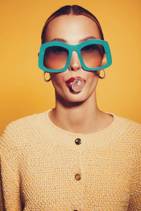 Young woman with oversized turquoise sunglasses blows a bubblegum bubble. She wears a yellow knitted sweater and gold hoop earrings on a bright yellow background. Fun and playful concept.