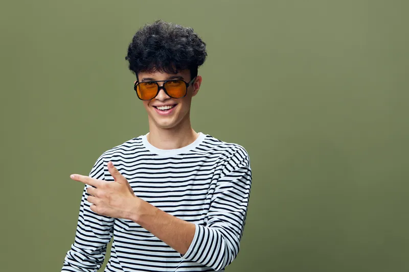 young man wearing sunglasses and striped shirt smiling and pointing with finger against green background, casual style and cheerful mood in studio lighting