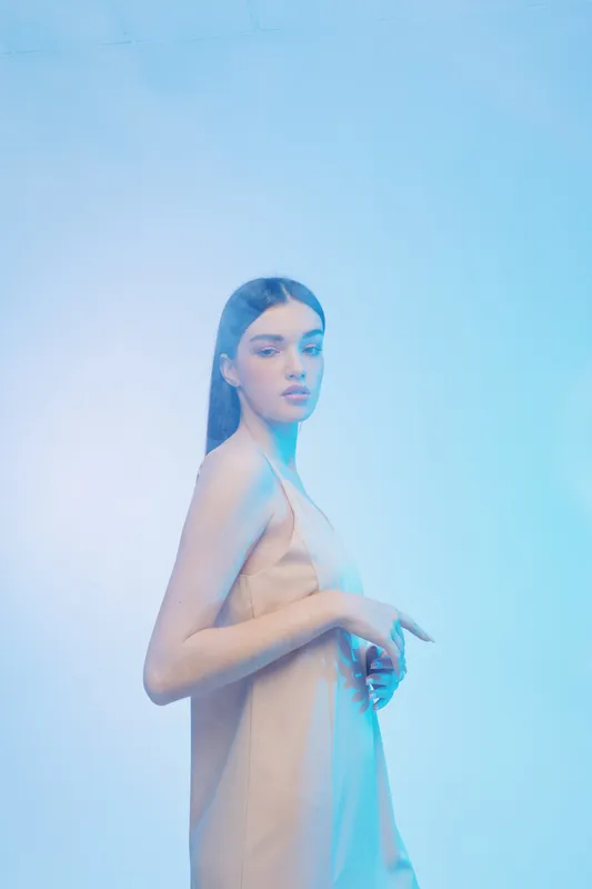 Young woman in a beige dress poses confidently against a soft blue background. The image highlights beauty, fashion, and elegance with minimalist styling and cool lighting.