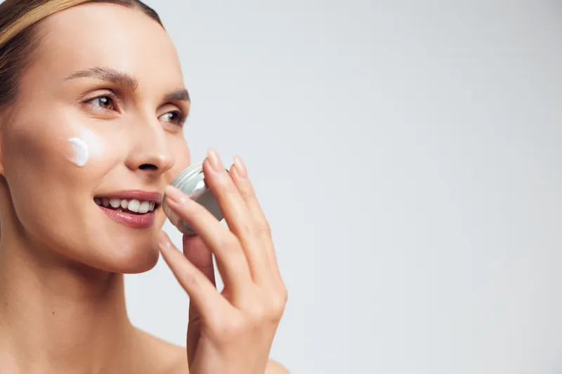 A smiling woman applies cream on her cheek using a small jar in a bright studio. The close-up shows skincare routine and natural beauty care concept with soft lighting.