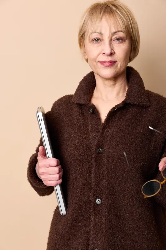 Senior woman portrait, confident business professional holding a tablet, smiling in studio against a beige background, warm jacket, calm expression. Modern technology in aging.