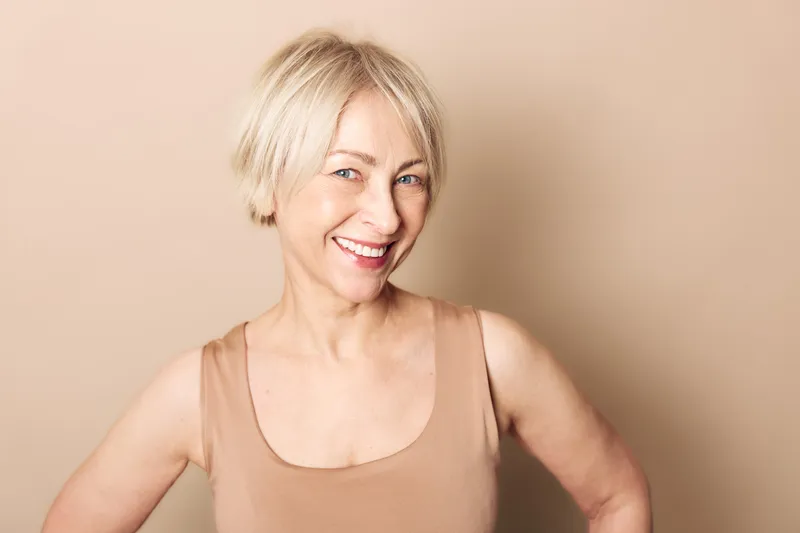 Smiling mature woman with short blonde hair wearing sleeveless top, posing confidently against beige background. Concept of happiness, aging, wellness, and natural beauty.