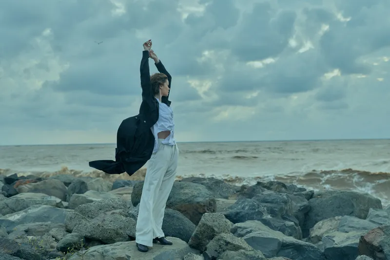 Young adult woman in white trousers and a black jacket stands on a windswept rocky shoreline, arms raised, ocean breeze fluttering fabric, a fashion moment by the sea
