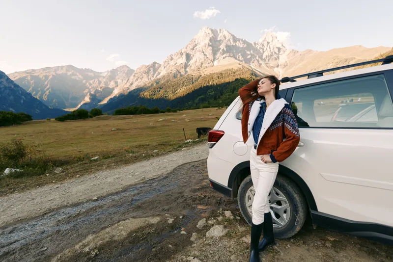 Woman leaning on white car beside mountain landscape and gravel road, travel adventure concept with stylish jacket and boots, enjoying scenic view and outdoor freedom.