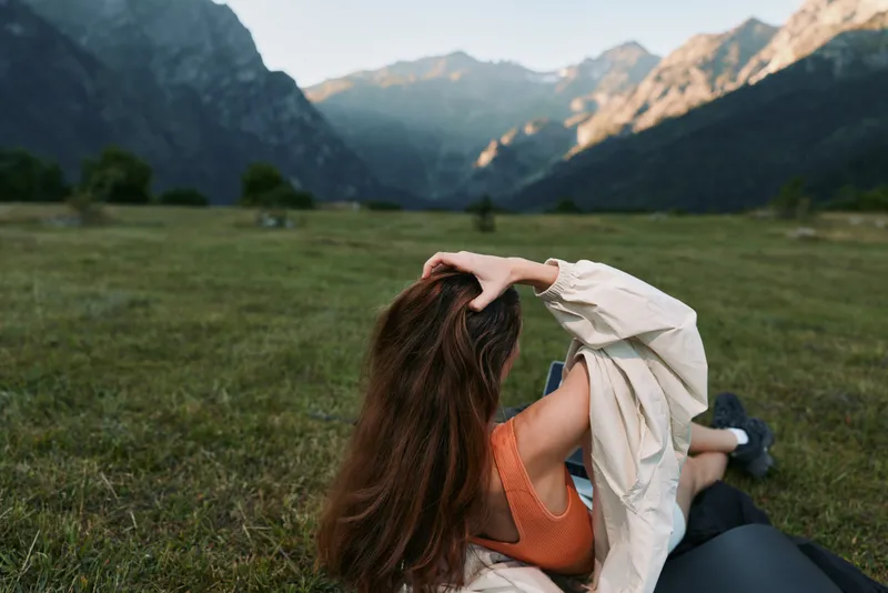 A person lies on a grassy meadow with towering mountain peaks in the background, enjoying the outdoors. The scene conveys relaxation, freedom, and a peaceful escape.