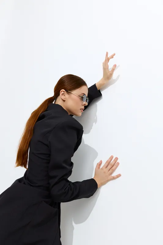Fashion woman with long brown hair wearing black outfit and sunglasses poses against a white wall, showcasing modern style and minimalistic elegance in bright lighting.