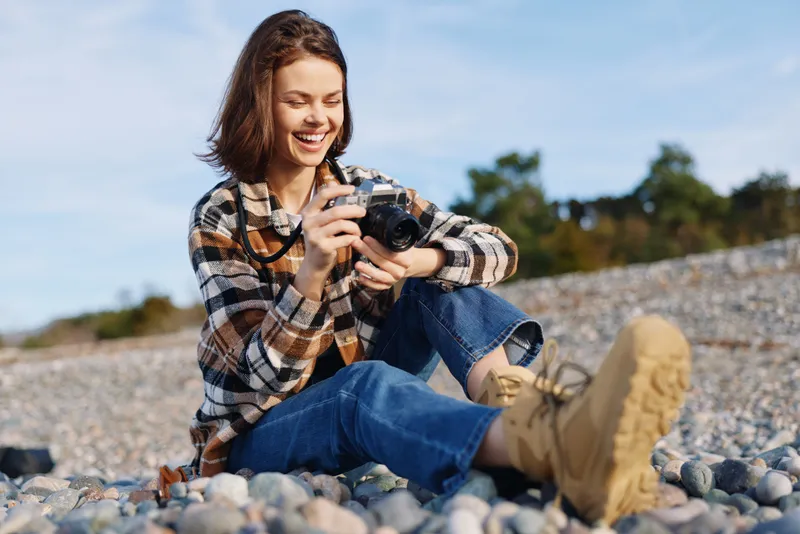 Photography, photographer, camera, outdoors, woman, casual, rocks and shoreline, joyful moment capturing scenery with lens on a pebble beach under a sunny blue sky and a light breeze.