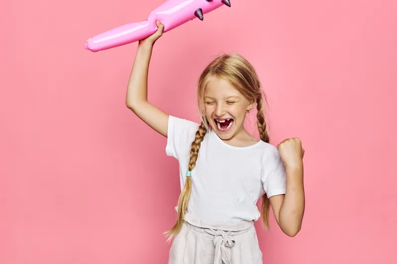Happy girl with long blond hair in pigtails celebrating with pink inflatable toy on pink background. She is smiling with closed eyes and raised fist in excitement.