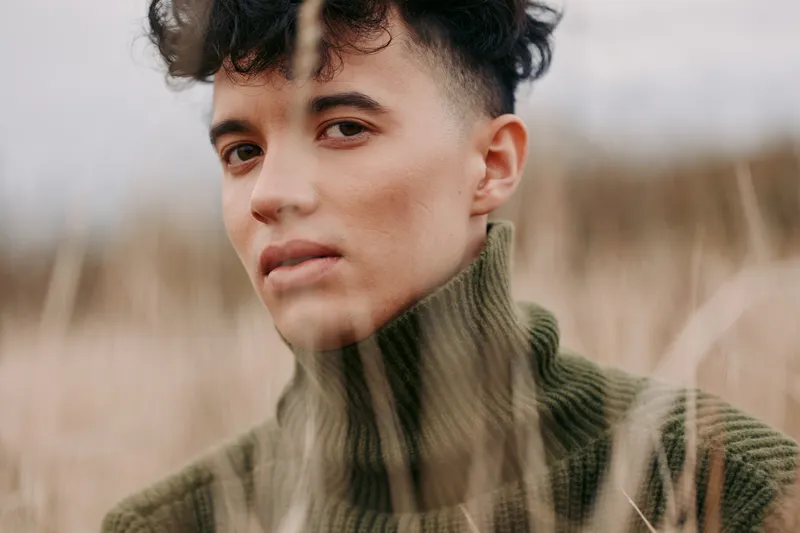 Close up portrait of a young person in a warm green knit turtleneck among tall dry grasses, an authentic, credible gaze with genuine expression conveying quiet confidence and real emotion.