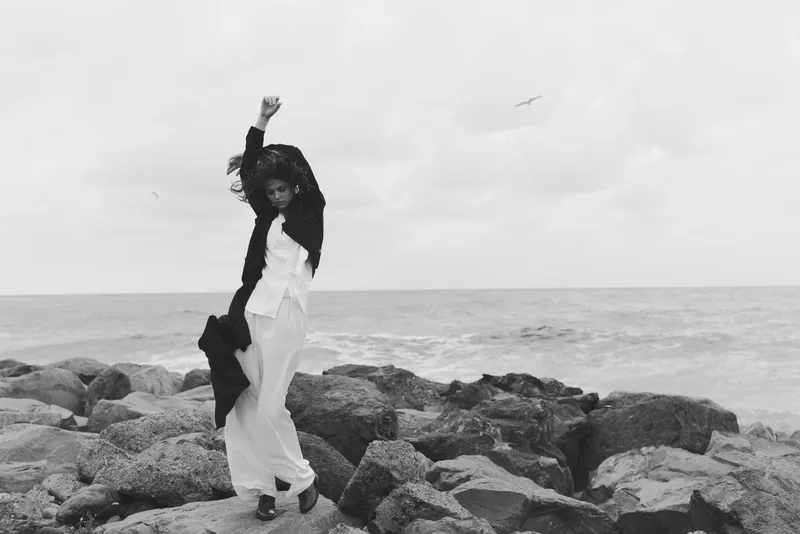 Fashion movement on rockyshore in monochrome seacape, wind tousling dress and scarf, dramatic silhouette by the ocean coast in soft light and spray of waves