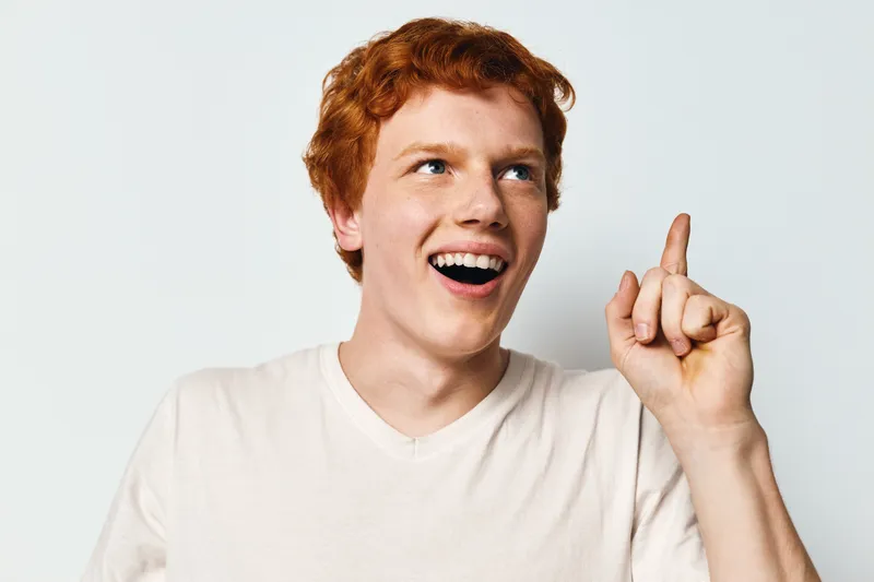 Young man with red hair and white shirt smiling and pointing up with finger on light background. Expression of idea, inspiration or realization with open mouth and blue eyes.