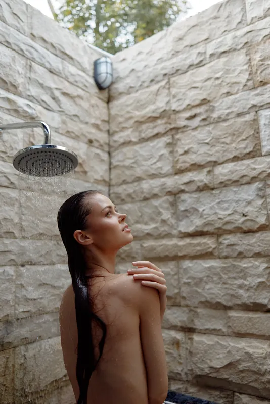 woman showering outdoors under rain showerhead with stone walls, natural light highlighting wet skin and serene expression in relaxing bath scene