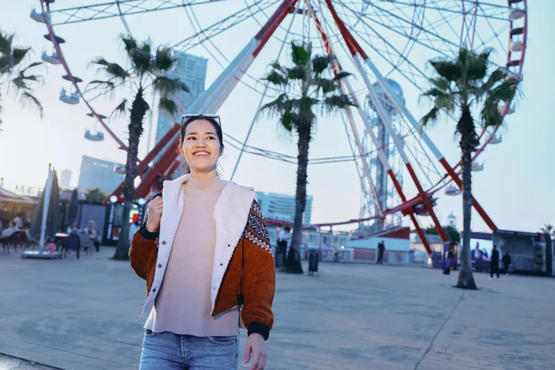 Smiling woman in casual jacket on a seaside boardwalk near a large ferris wheel, daytime outdoor scene, travel and leisure vibe, candid moment of joy, fashion style against an urban waterfront